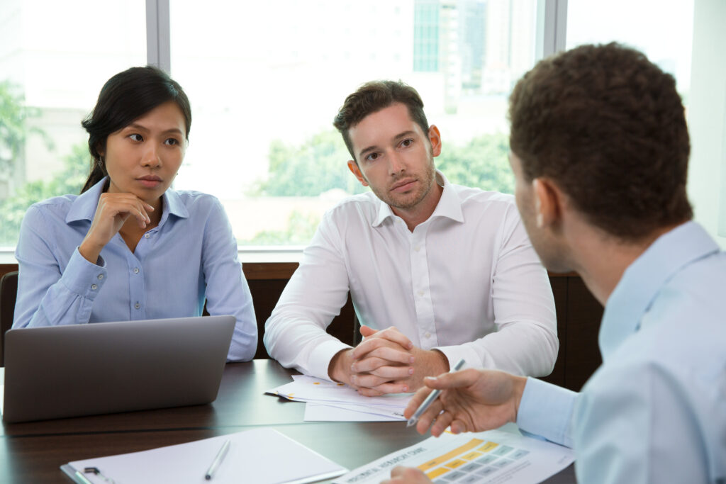 Young businessman and businesswoman sitting at a desk with an office window in the background, listening to a male colleague, representing continued learning in business.